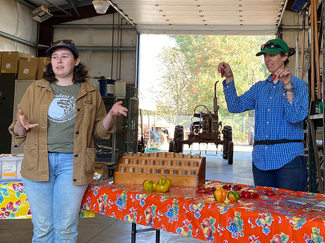 Two young women stand at a table, one of them holding up green jalapeno peppers, and the other one talking.